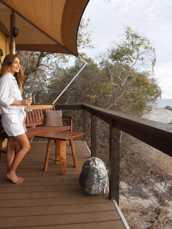 A girl stands with a glass of wine in hand, admiring the beautiful Cygnet Bay and remote location that surrounds her Glamping Safari Tent in the Diver's Village of Cygnet Bay Pearl Farm.
