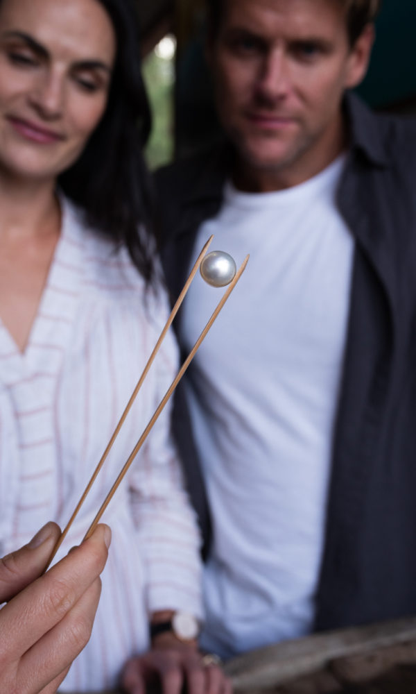Between a pair of pearl tongs, a tour guide holds a beautiful white Australian South Sea pearl during a Pearl Grading Tour. The guests in attendance look on in amazement.
