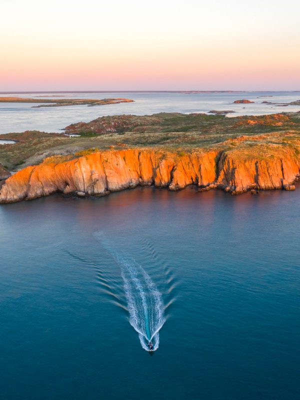 A boat travels on the serene, calm waters of the Kimberley sea towards Cygnet Bay Pearl Farm. In the background is an island, part of the Archipelago of islands, glowing in orange hues from the stunning sunset. The sunset lights up the sky in a warm gradient, in contrast with the azure waters in the foreground.