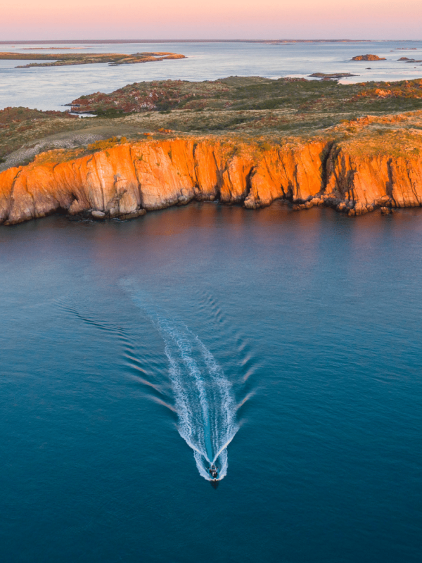 A boat travels on the serene, calm waters of the Kimberley sea towards Cygnet Bay Pearl Farm. In the background is an island, part of the Archipelago of islands, glowing in orange hues from the stunning sunset. The sunset lights up the sky in a warm gradient, in contrast with the azure waters in the foreground.