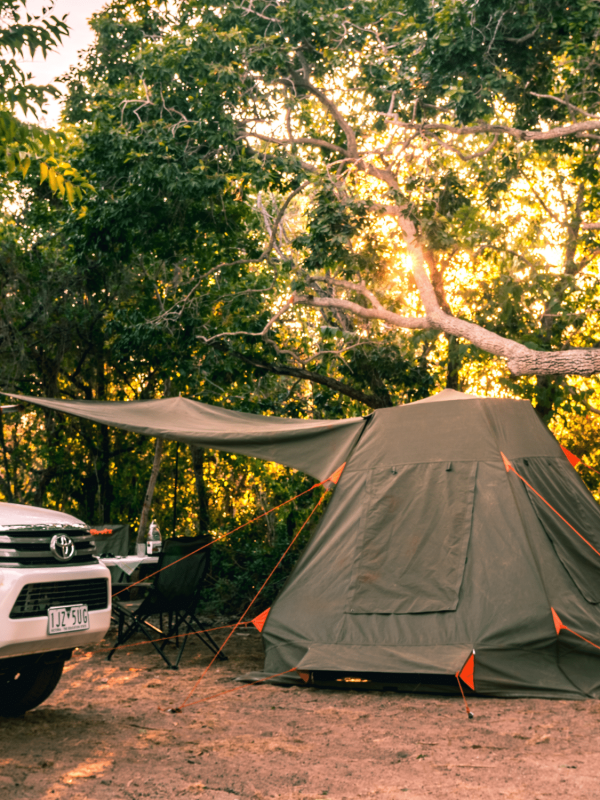 A 4WD sits at a Diver's Creek campsite, with a tent pitched on the rugged terrain. The tent sits under a tent with green foliage and is surrounded by Australian native shrubs to create privacy for campers.