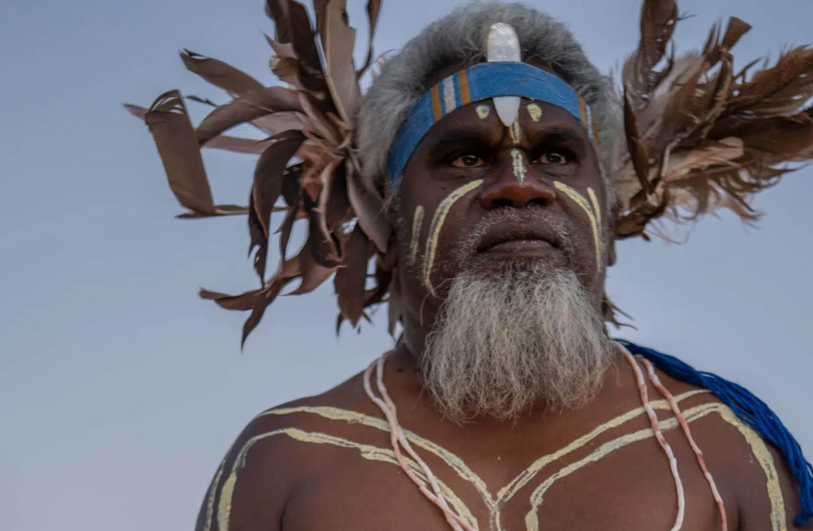 A Bardi Jawi man is dressed in traditional clothing, with culturally significant body paint covering his face, chest and arms. He wears a headband with a piece of Mother of Pearl resting between his forehead and the headband, shining with great lustre, and at the back of his head are several feathers that flourish from the sides of his head. He holds a boomerage in both hands and stares into the distance.