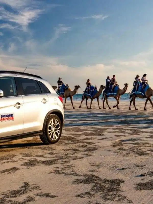 A 4WD sits on a white sandy Broome beach, with the waves crashing in the background and a line of camels with tourists sitting on them, enjoying Cable Beach.