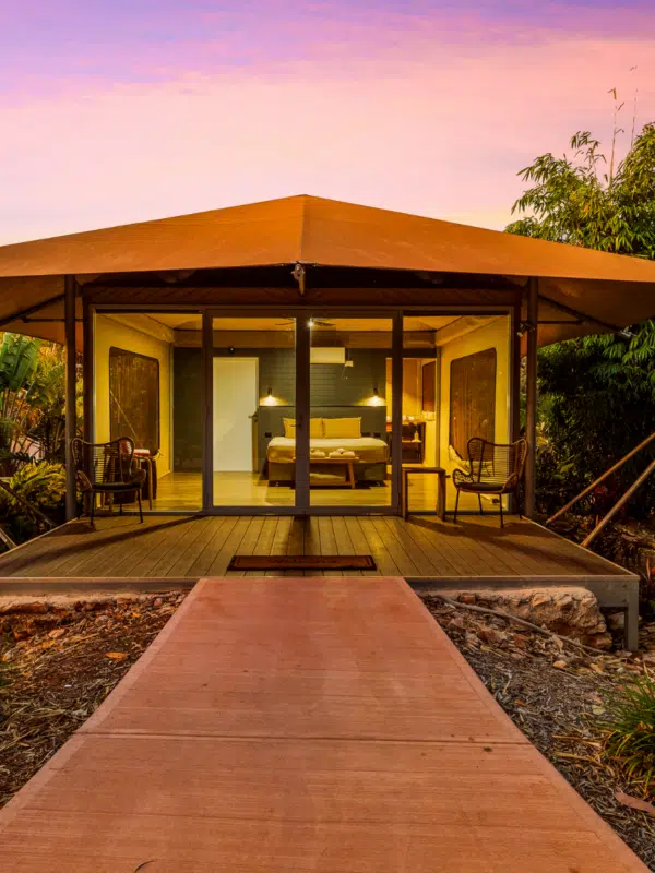 A path leads to a Pearler's Village Safari Tent glowing from within at dusk. At this time of the day you can see inside the tent, showing the comfortable king bed with two ambient lamps on either side of the bed, the door to the ensuite of the tent and the passage to the self countained kitchenette area as well. Behind the tent is a beautiful Kimberley sunset, with pink and purple hues, and surrounding the tent are native, tropical plants - creating serenity and calm for those staying in this accommodation.