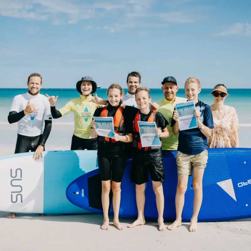 A group of adults and children stand on a Broome beach by a surfboard, smiling and enjoying the seaside. The children hold a certificate stating Congratulations that they have participated in an Ocean Heroes event.