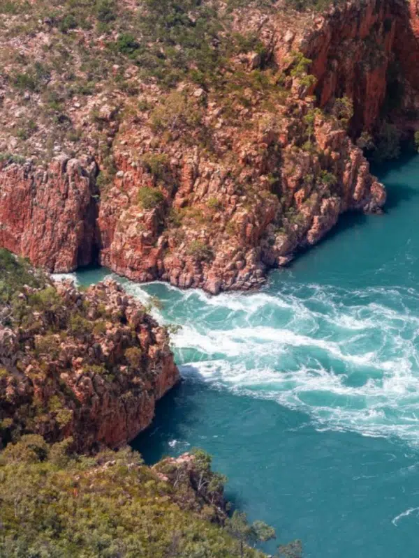 An aerial visual of the intense, rushing waters of the natural phenomenon that is the Horizontal Falls. This marine marvel occurs due to the tides pushing through the two islands, side by side, and produce a 'waterfall' through the landmarks. The sea is turqouise blue, and aside from the tidal waters creating white foaming waves, the rest of the ocean is calm and still.