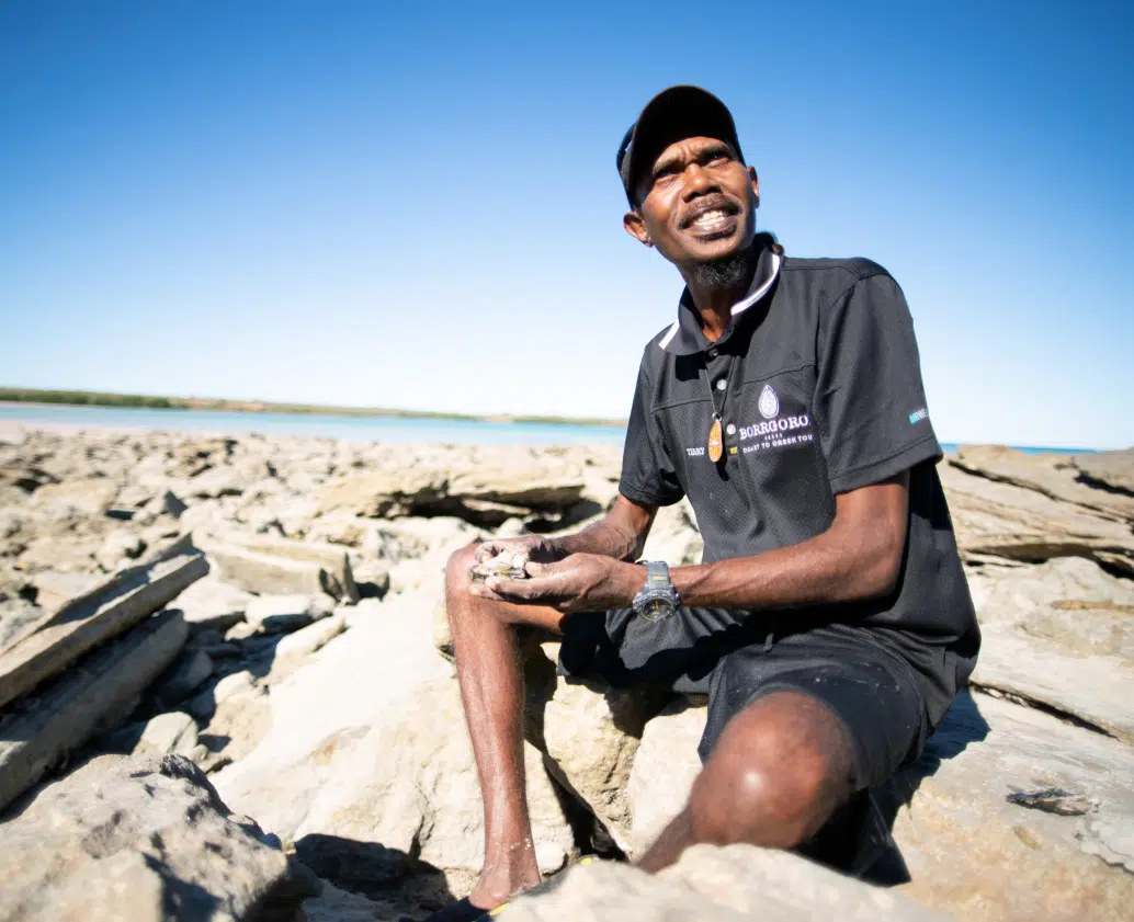 Terry Hunter sits on a beach during his Borrgoron Cultural Tour. Behind him is the shoreline of Cygnet Bay.