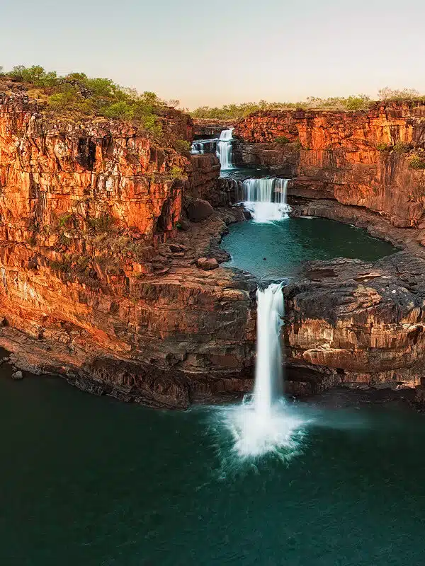 A stunning visual of Mitchell Falls, an iconic Kimberley landmark, that shows the red rocks lit up during sunset and the streaming waterfall cascading across these rocks.