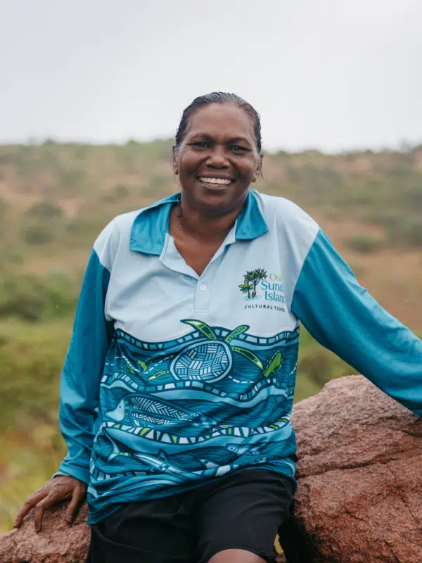 Rosanna Angus sits on a rock, wearing her Oolin Sunday Island Cultural Tours shirt. She smiles wide, looking at the camera, while in the background is a large expanse of native Australian shrubs and trees.