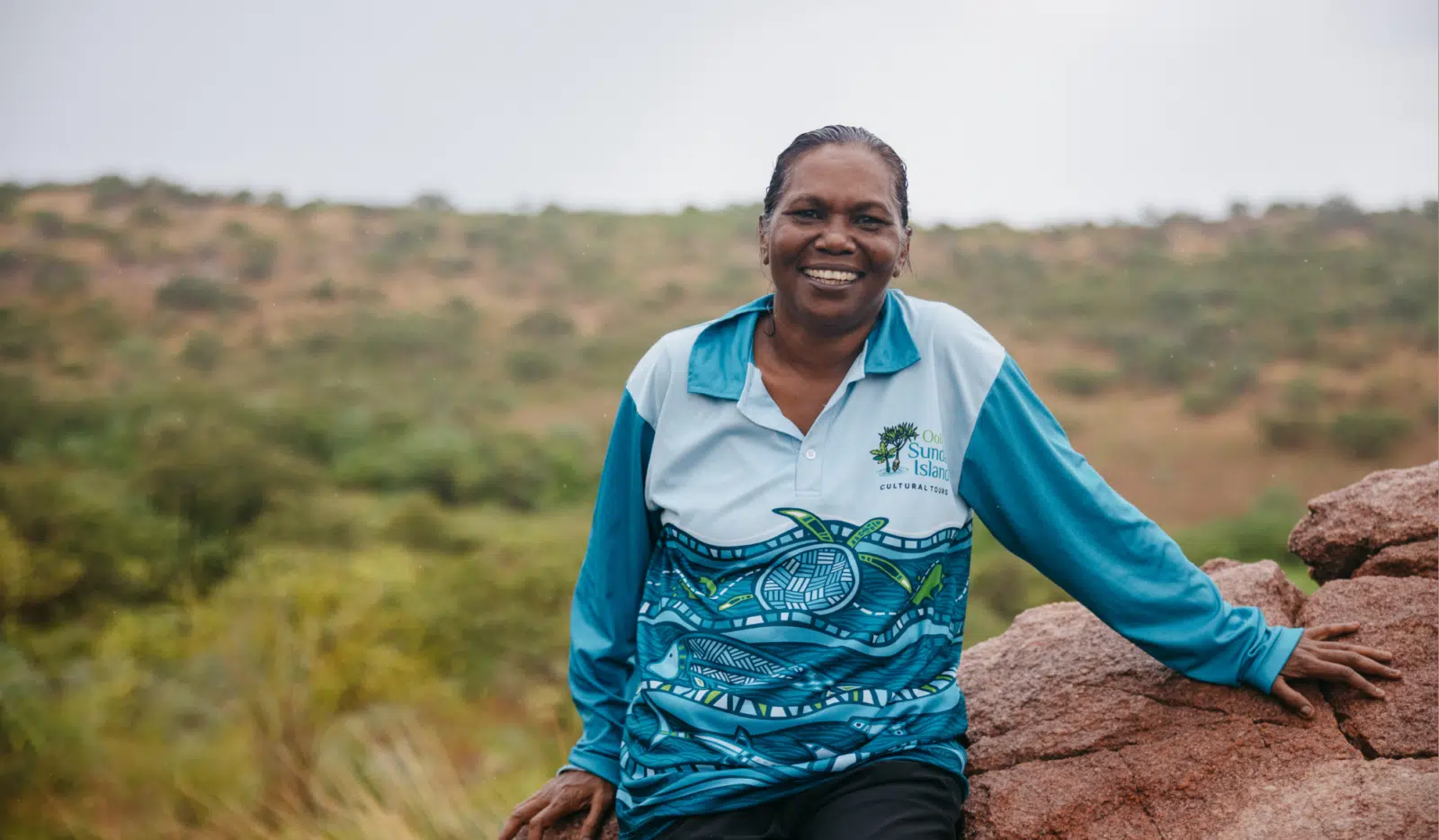 Rosanna Angus sits on a rock, wearing her Oolin Sunday Island Cultural Tours shirt. She smiles wide, looking at the camera, while in the background is a large expanse of native Australian shrubs and trees.