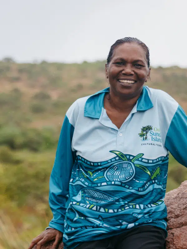 Rosanna Angus sits on a rock, wearing her Oolin Sunday Island Cultural Tours shirt. She smiles wide, looking at the camera, while in the background is a large expanse of native Australian shrubs and trees.