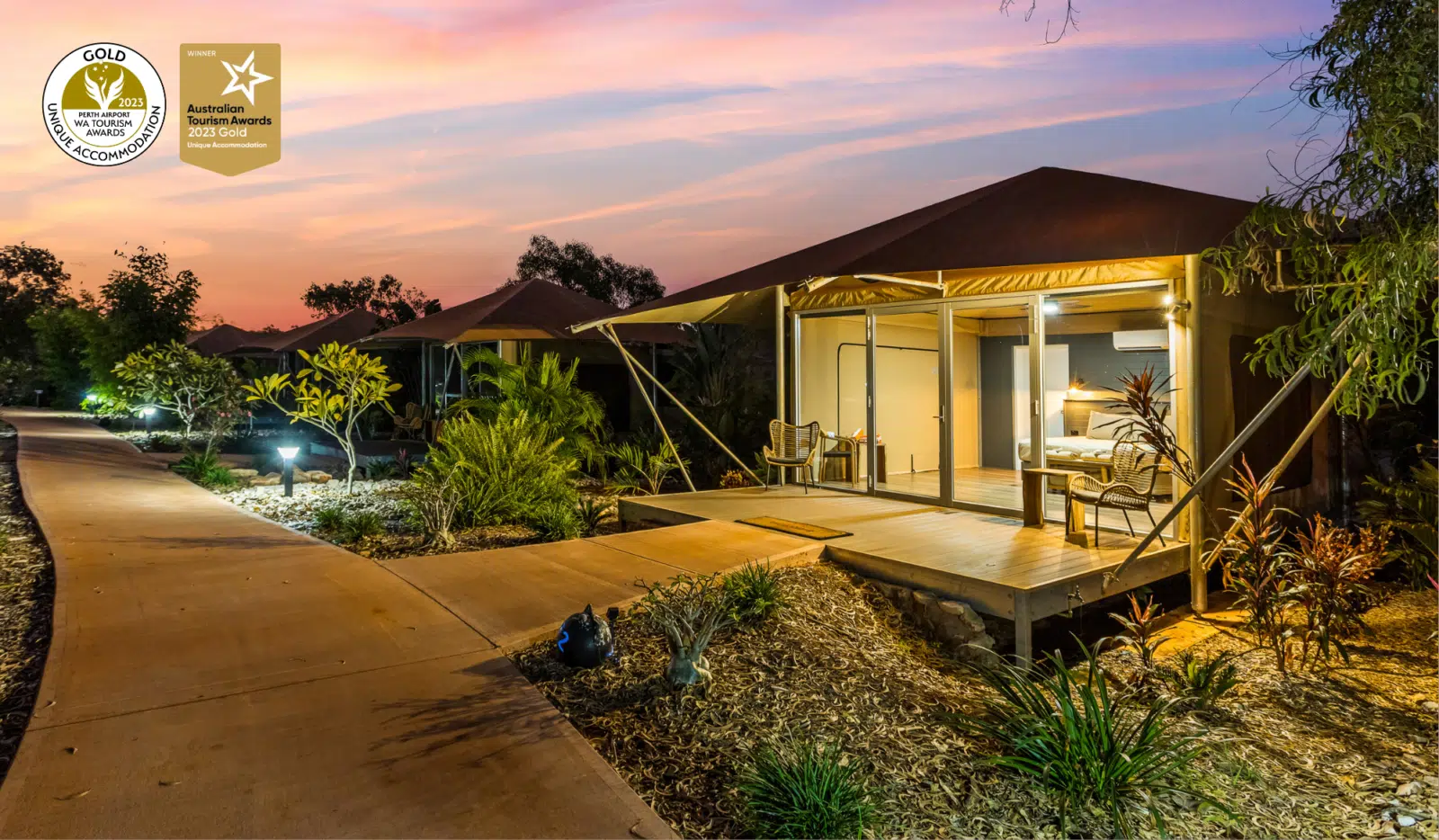A path leads to a Pearler's Village Safari Tent glowing from within at dusk. At this time of the day you can see inside the tent, showing the comfortable king bed with two ambient lamps on either side of the bed, the door to the ensuite of the tent and the passage to the self countained kitchenette area as well. Behind the tent is a beautiful Kimberley sunset, with pink and purple hues, and surrounding the tent are native, tropical plants - creating serenity and calm for those staying in this accommodation.