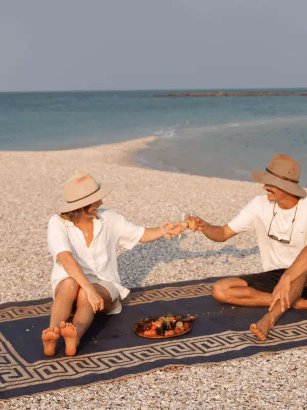 A group of friends share snacks on a rug with a beautiful, glowing sunset in the distance. They sit on an island of shells, a secret beach, that shifts in size depending on the tidal movements known to the Dampier Peninsula.