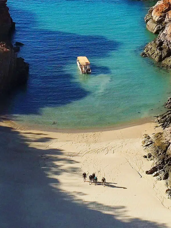 A group of people walk onto shore, after being on an Oolin Sunday Island Explorer tour with Rosanna Angus, ready to explore and learn about the stunning Kimberley region. In the background is the boat they travelled on, which is anchored in turquoise waters.