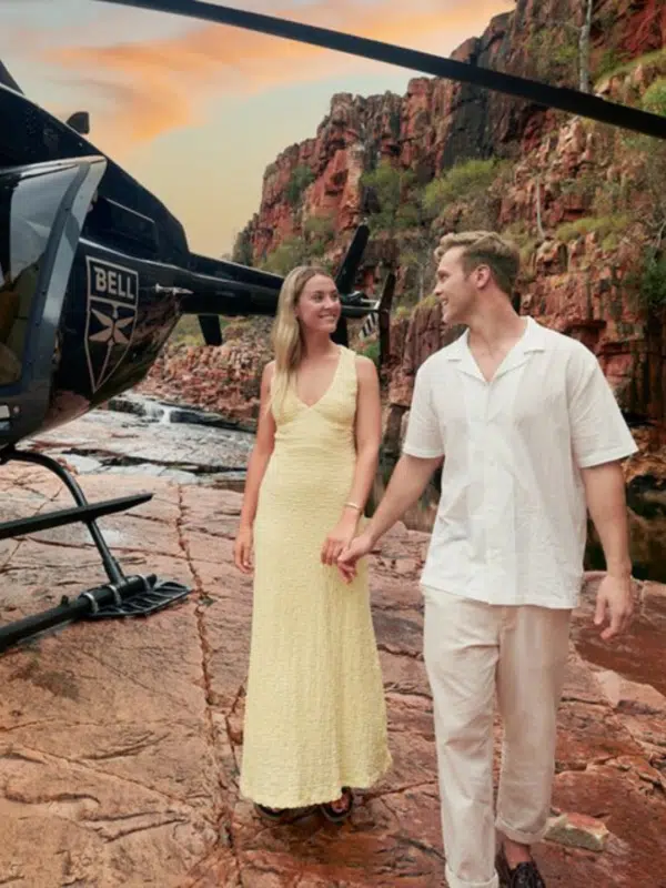 A couple walk along a Kimberley gorge, smiling at each other as they enjoy the beautiful sunset surrounding them. In the distance are red, rocky cliffs, waterfalls, and next to them is a helicopter that transported them to this special, secret spot during their Ventur Atlas Tour.