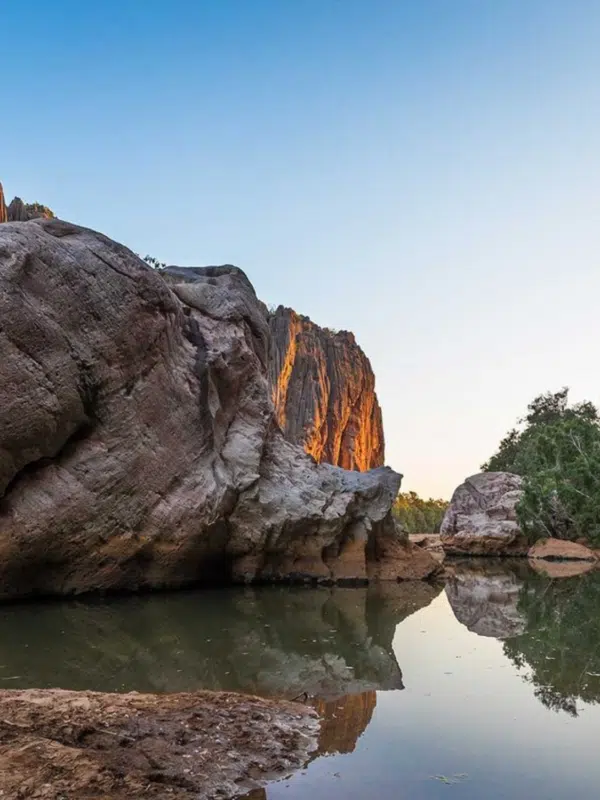 A beautiful photo of a Kimberley gorge at sunset, with the golden light basking the red rock. In the foreground the gorge cliffs are reflected in the tranquil waters of a lagoon.
